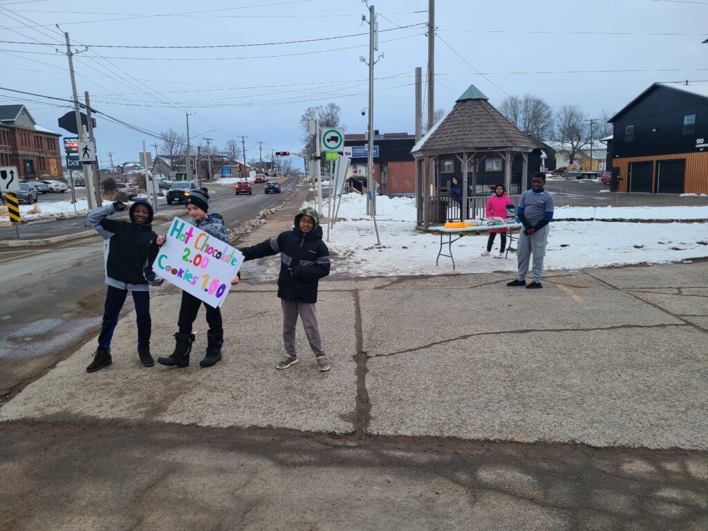 Junior youth hold up a sign for cars that advertises their hot chocolate sale. Through this project, the junior youth aimed to raise money for a new ping pong table for their neighbourhood centre.